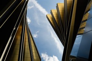 Upward view between modern gold‑toned building structures with blue sky and clouds above.