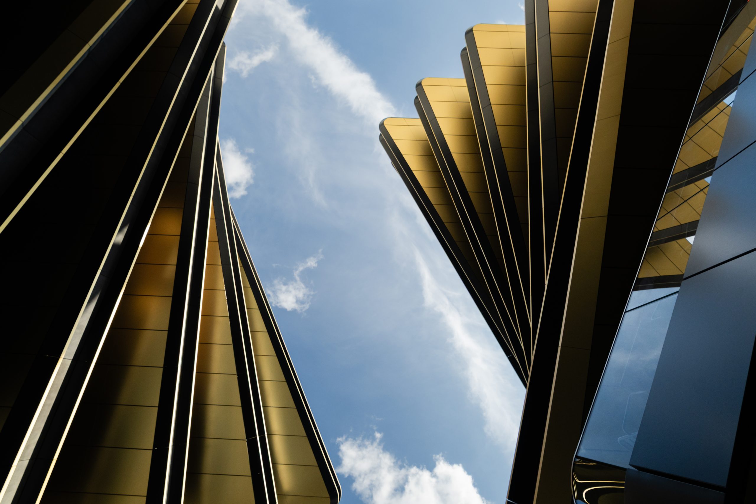 Upward view between modern gold‑toned building structures with blue sky and clouds above.