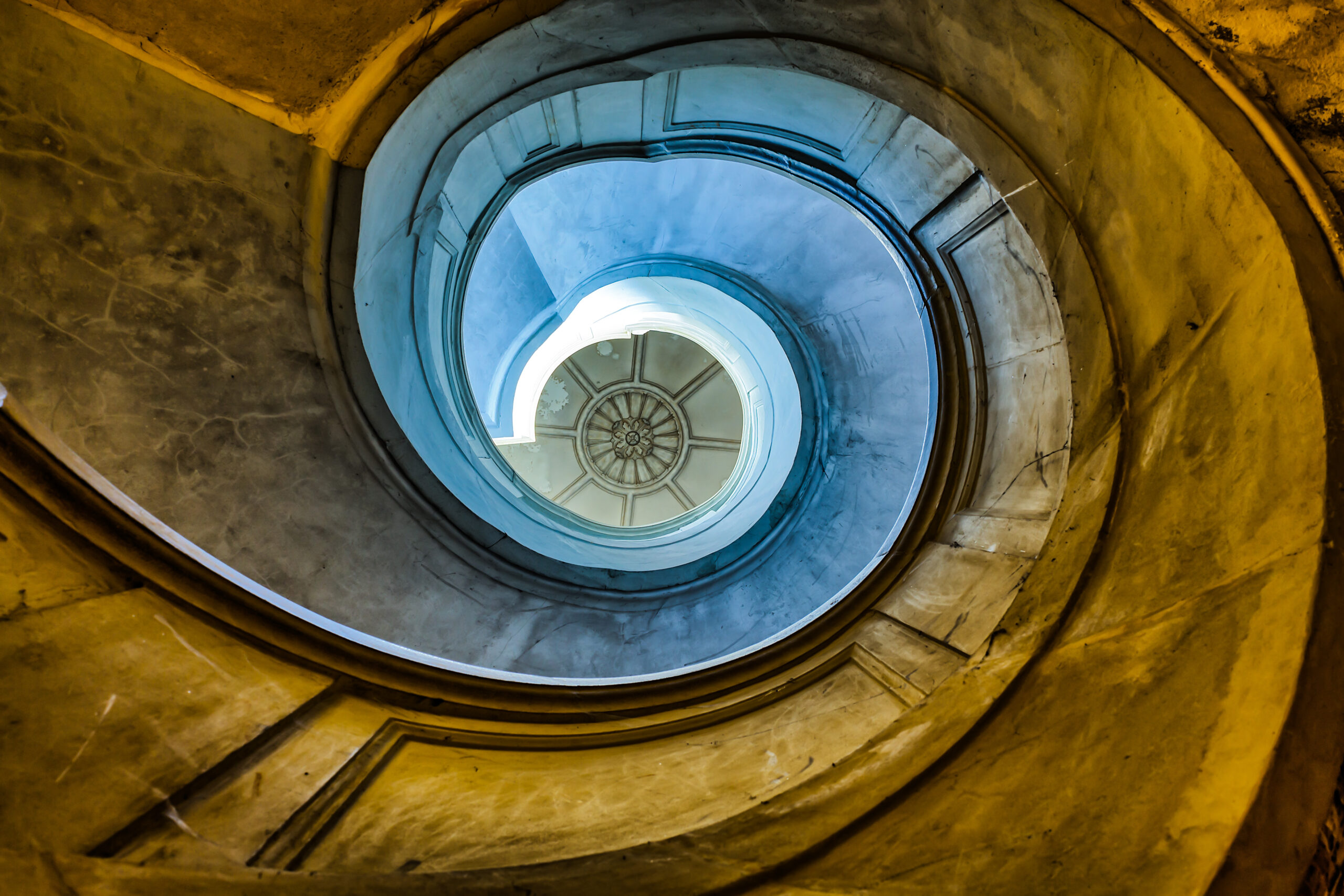 An architectural spiral staircase viewed from below, curving towards a circular skylight at the centre.